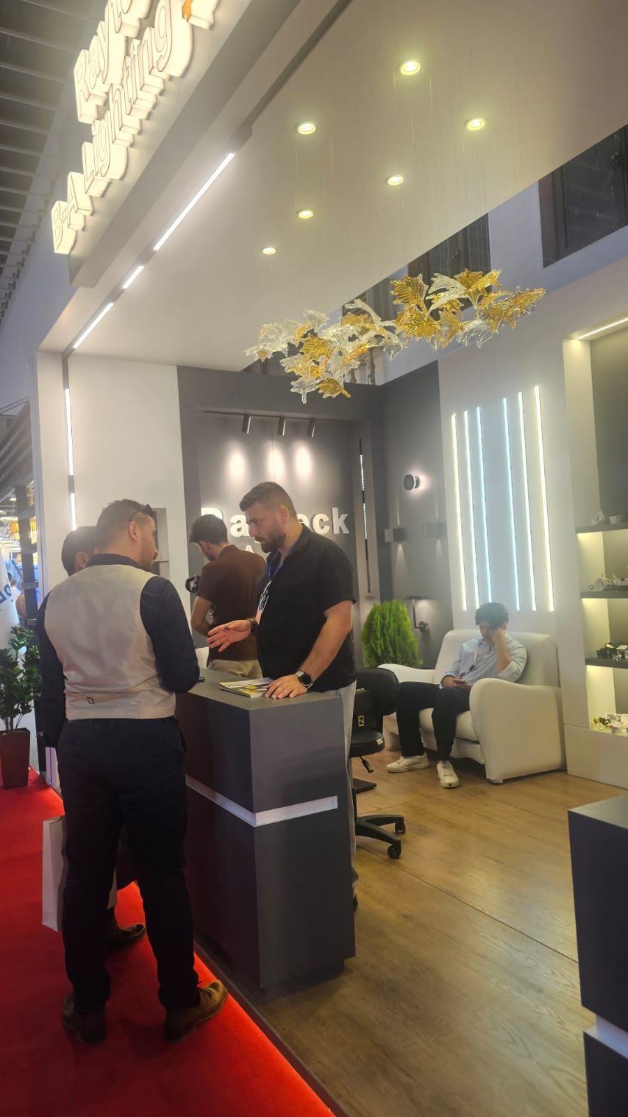 The RAYTECK stand at BUILDEX 2026, its Rayteck Lighting sign lit above a grey reception desk, with a decorative glass-leaf chandelier overhead.