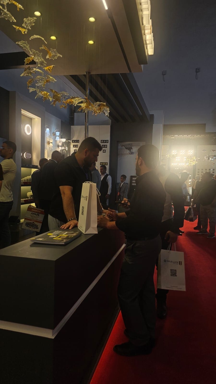 Visitors at the RAYTECK stand, brochures and a catalogue bag on the reception desk, product walls lit behind them.